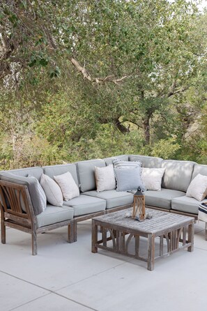 A grey sectional sofa with patterned cushions, a wooden bench, and a small table with a lantern, set outdoors with trees in the background. - Private 14 Acre Estate on Barton Creek with Two Homes, Pool and Multiple Gardens (Austin)