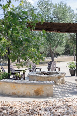 An outdoor seating area with wooden chairs and tables, a stone fire pit, and a pergola overhead. - Private 14 Acre Estate on Barton Creek with Two Homes, Pool and Multiple Gardens (Austin)