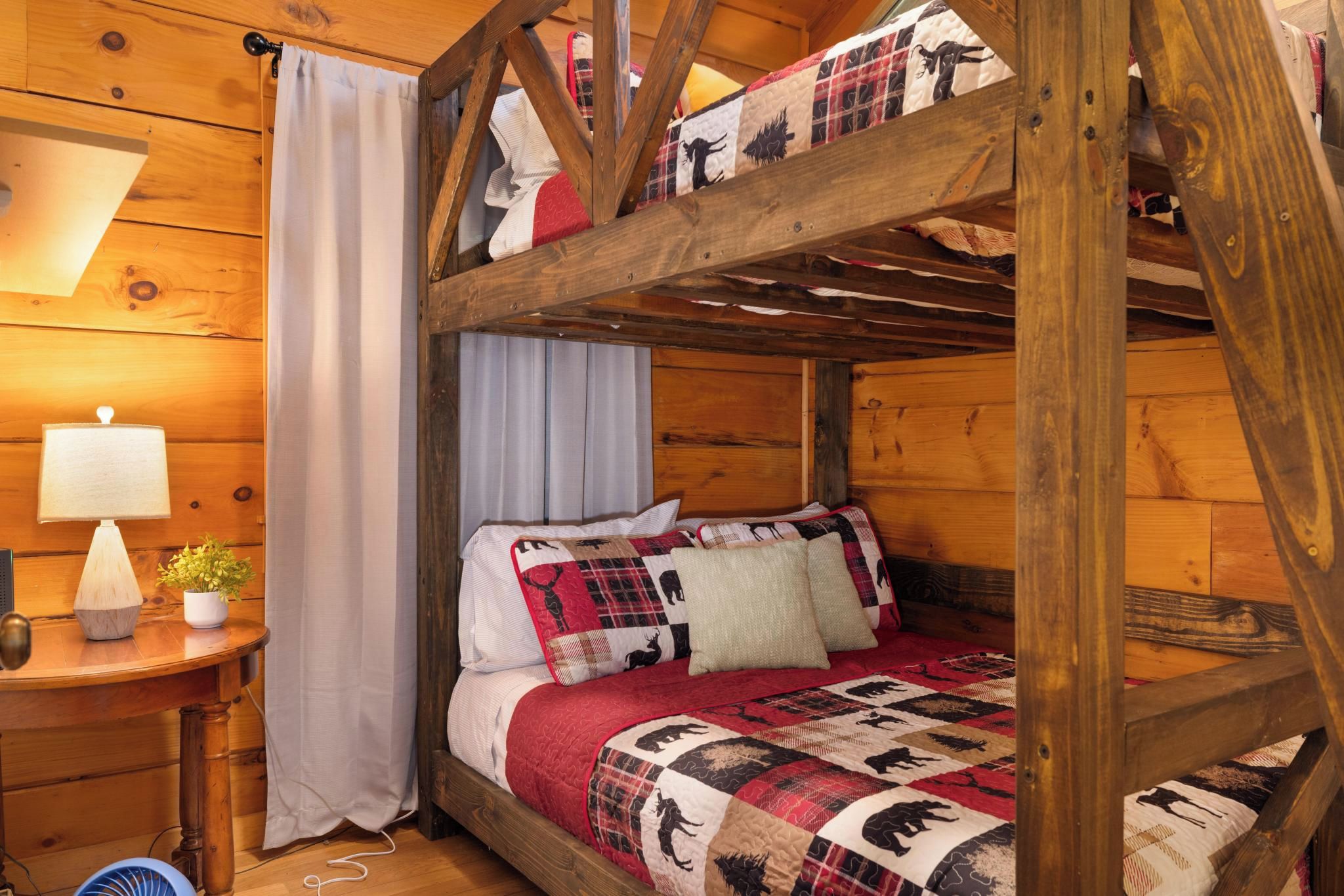 Una cama litera con colcha roja y negra, una mesita de noche con lámpara y una ventana con cortinas.