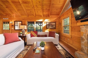 Ein gemütliches Blockhaus-Interieur mit Holzbalkendecke, einem weißen Sofa, einem Couchtisch mit Pflanze und einem an der Wand befestigten Fernseher.