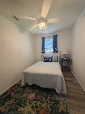 Ein Schlafzimmer mit einem Bett, einem Deckenventilator, einem Fenster mit Vorhängen, einem Nachttisch mit Lampe und einem Teppich mit Sternfischmuster.