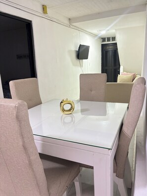 Una habitación con una mesa de vidrio, sillas color beige, un adorno dorado, una puerta negra y un televisor montado en la pared.