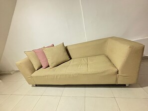 Un sofá color beige con dos almohadas frente a una pared lisa, sobre un piso de baldosas.