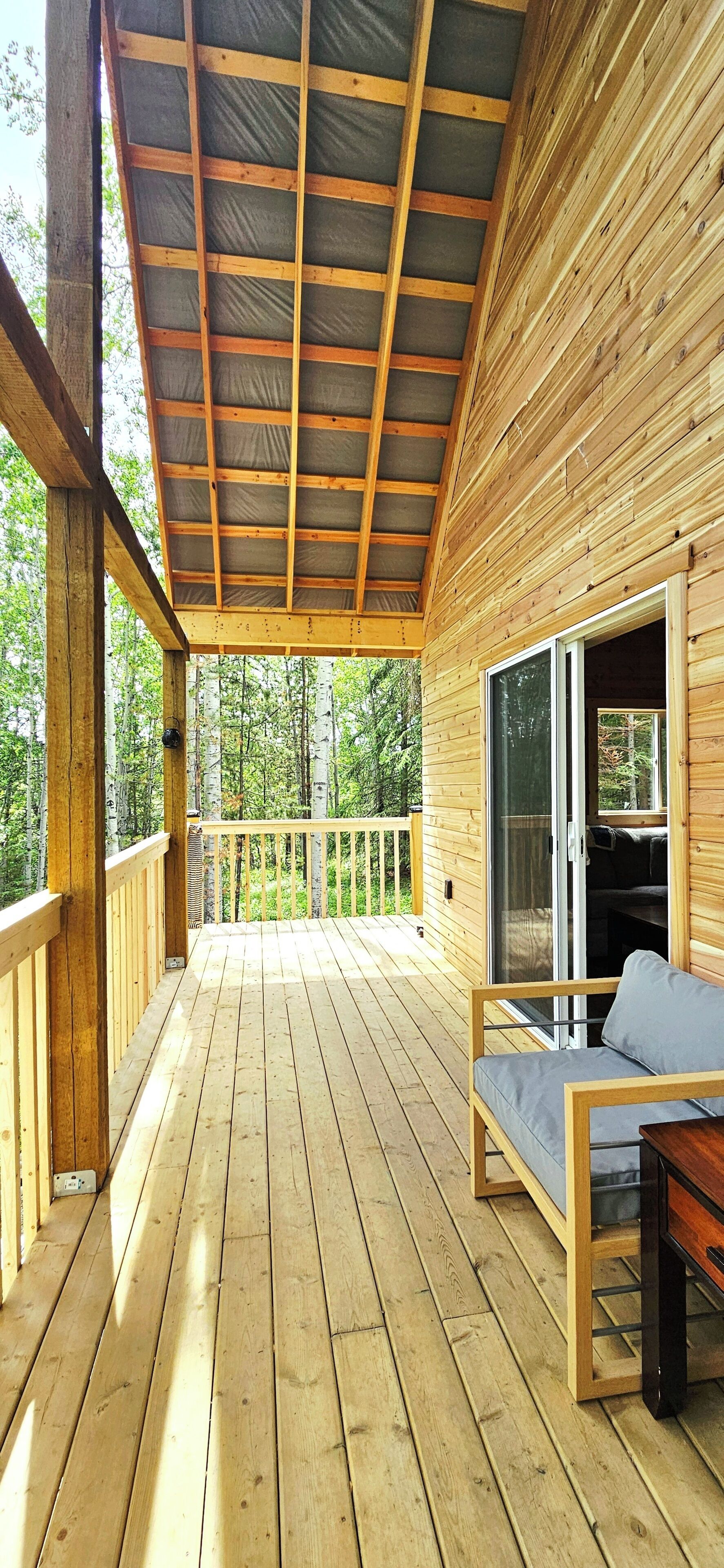 Eine Holzterrasse mit überdachtem Bereich, einer Sitzgelegenheit mit Kissen und Blick auf einen Wald.