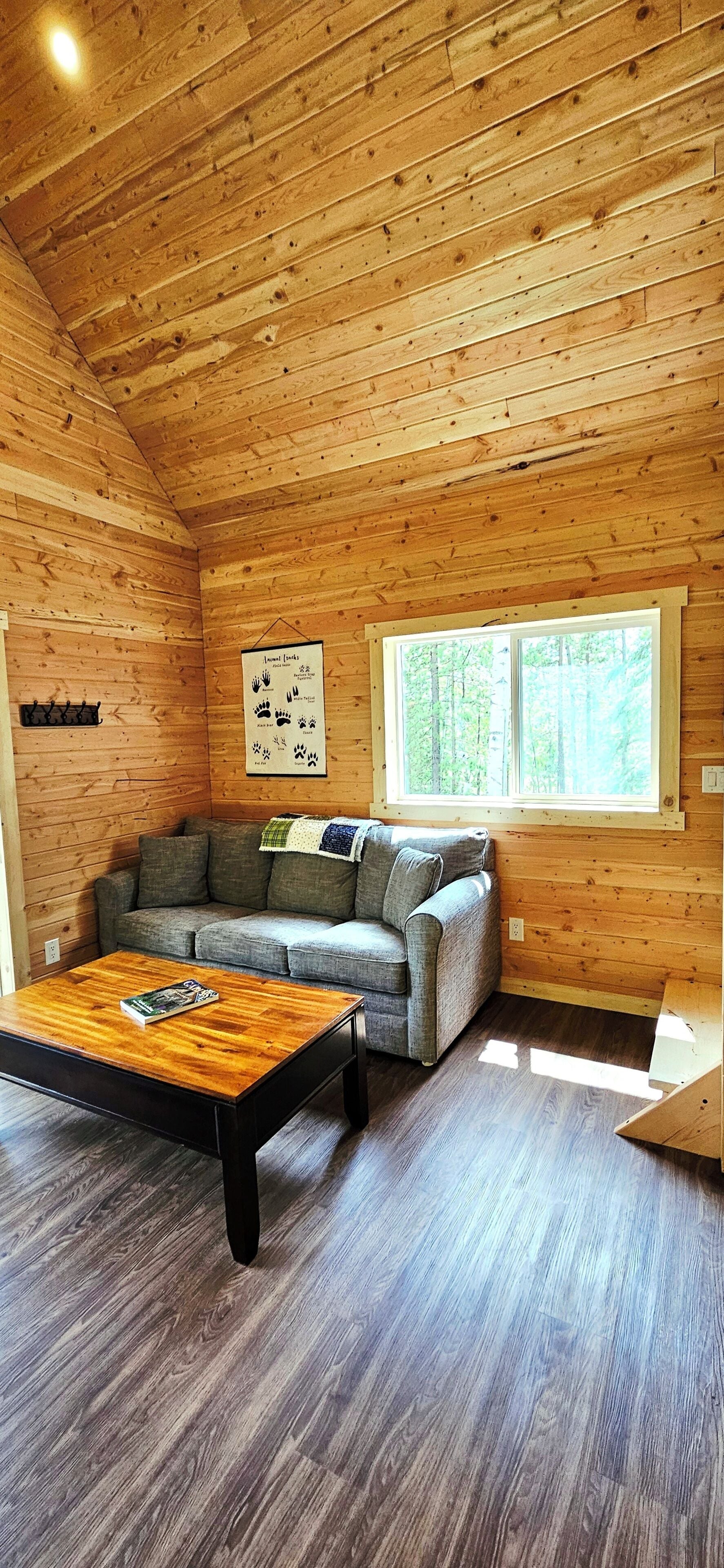 Ein gemütliches Cabin-Interieur mit Holzboden, einem grauen Sofa, einem Holztisch und einem Fenster mit Blick auf einen Wald.