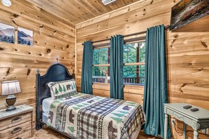 Quarto de cabana de madeira com cama, mesa de cabeceira, mesa e janela com cortinas.