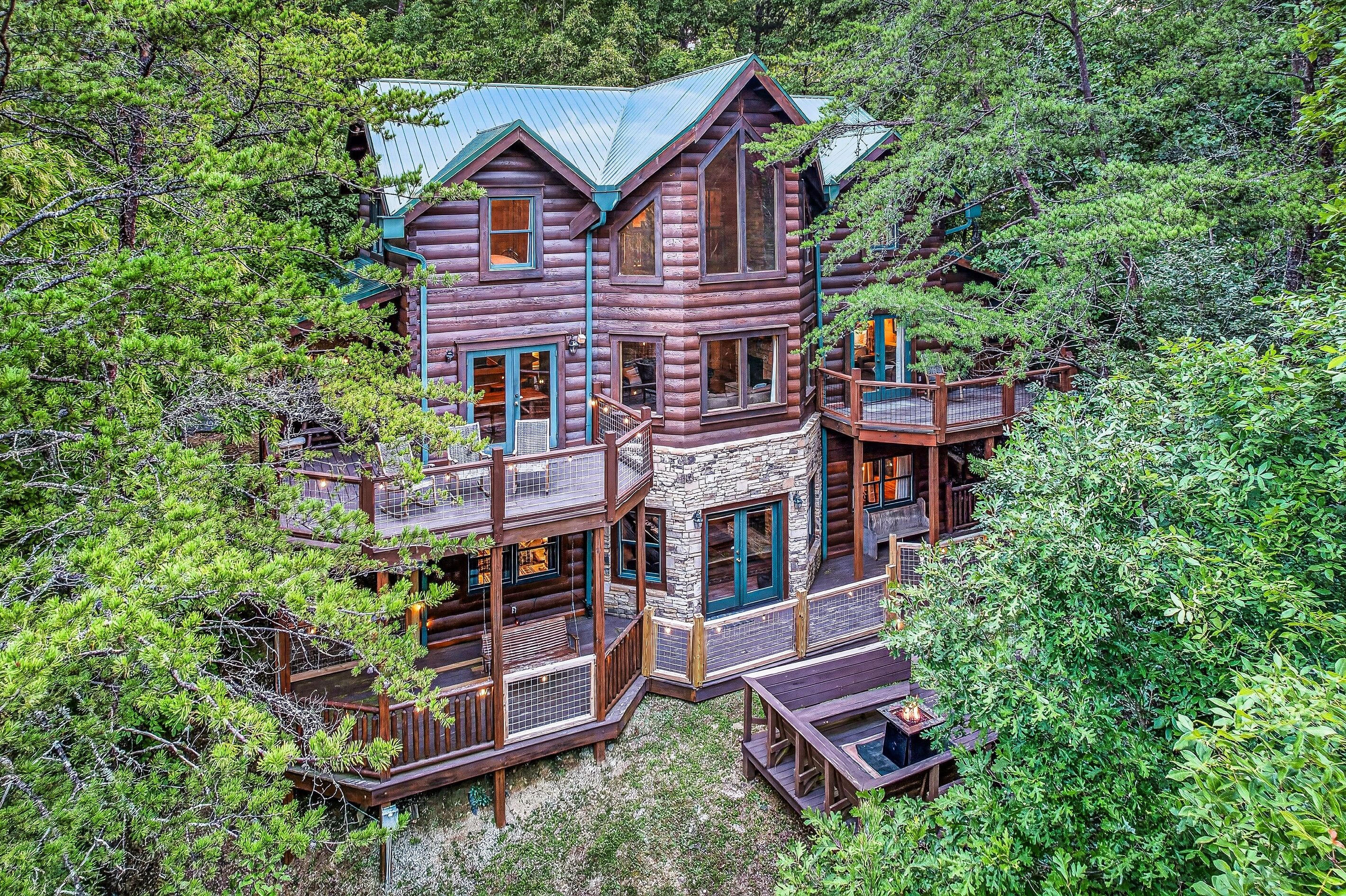 A multi-level log cabin with a balcony and a deck, surrounded by dense forest.