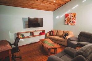 Een woonkamer met een wandmontage flatscreen televisie, een boekenplank, een beige bank, een grijze fauteuil en een houten salontafel.