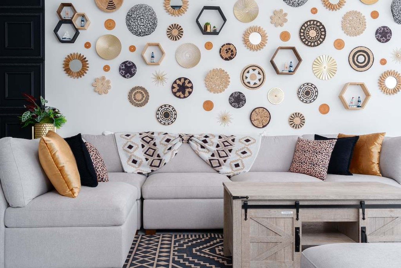 Ein modernes Wohnzimmer mit einem grauen Ecksofa, gemusterten Kissen, einem Couchtisch aus Holz und einer Wand, die mit verschiedenen dekorativen Tellern und Spiegeln geschmückt ist.