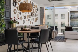 Een moderne woonkamer met een eethoek, met een grote geweven lamp, diverse kunstwerken aan de muur en uitzicht op buiten.