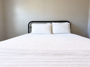 Un llit amb un edredó blanc i dos almoinos, un capçal metàl·lic i parets sense decoració.