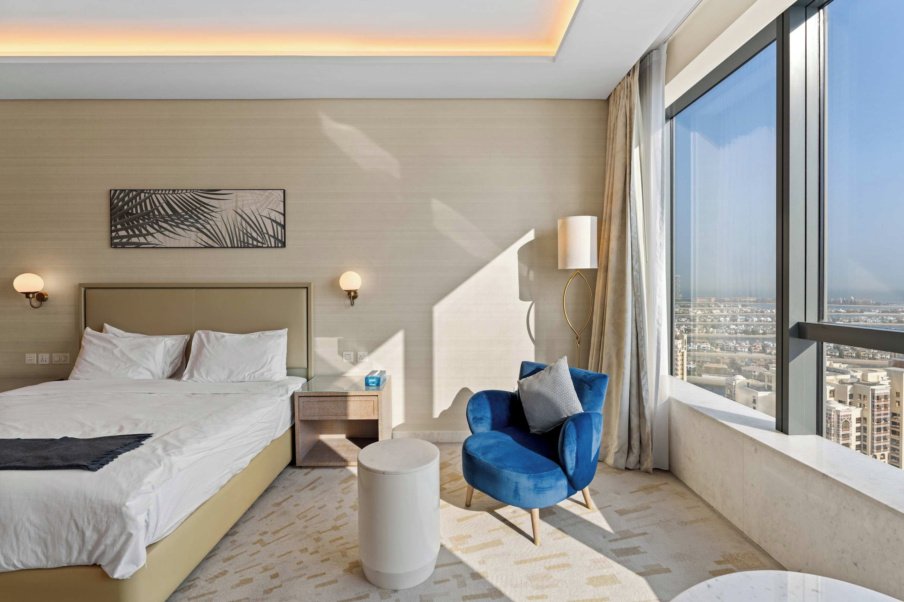 Ein Hotelzimmer mit einem großen Bett, einem blauen Sessel, einem runden Couchtisch und Blick auf die Stadt durch große Fenster.