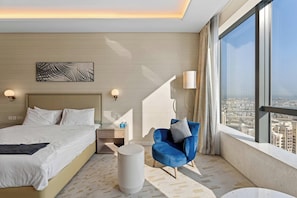 Ein Hotelzimmer mit einem großen Bett, einem blauen Sessel, einem runden Couchtisch und Blick auf die Stadt durch große Fenster.
