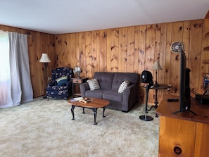 A living room with a wooden paneled wall, a grey sofa, a blue armchair, a coffee table, and a television. - Cozy 1-bedroom house in pleasant Guilford with AC (Guilford)