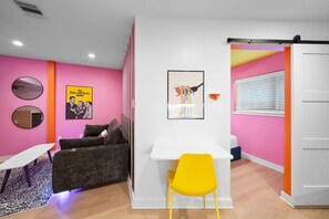Ein Zimmer mit einer rosafarbenen Wand, einem weißen Schreibtisch, einem gelben Stuhl, einem grauen Sofa und einem Plakat von 'The Dick Van Dyke Show'.
