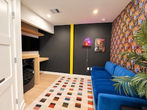 Ein modernes Wohnzimmer mit einem blauen Sofa, einer Wand mit Muster, einer Küchenzeile inklusive Waschmaschine und einem farbenfrohen Teppich.