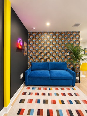 Ein Zimmer mit einem blauen Sofa, tapetiertem Wandhintergrund, einem Neon-Schild und einem farbenfrohen Teppich.