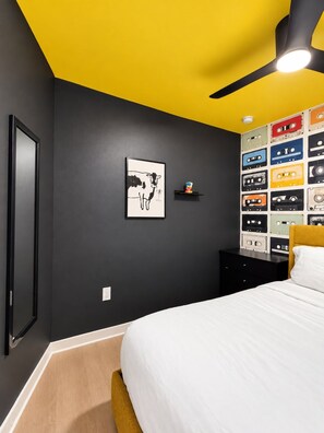 Ein Schlafzimmer mit schwarzer Wand, gelber Decke, einem Bett mit weißer Bettwäsche, einem schwarzen Nachttisch, einem gerahmten Bild und einer Wand voller farbiger Kassetten.