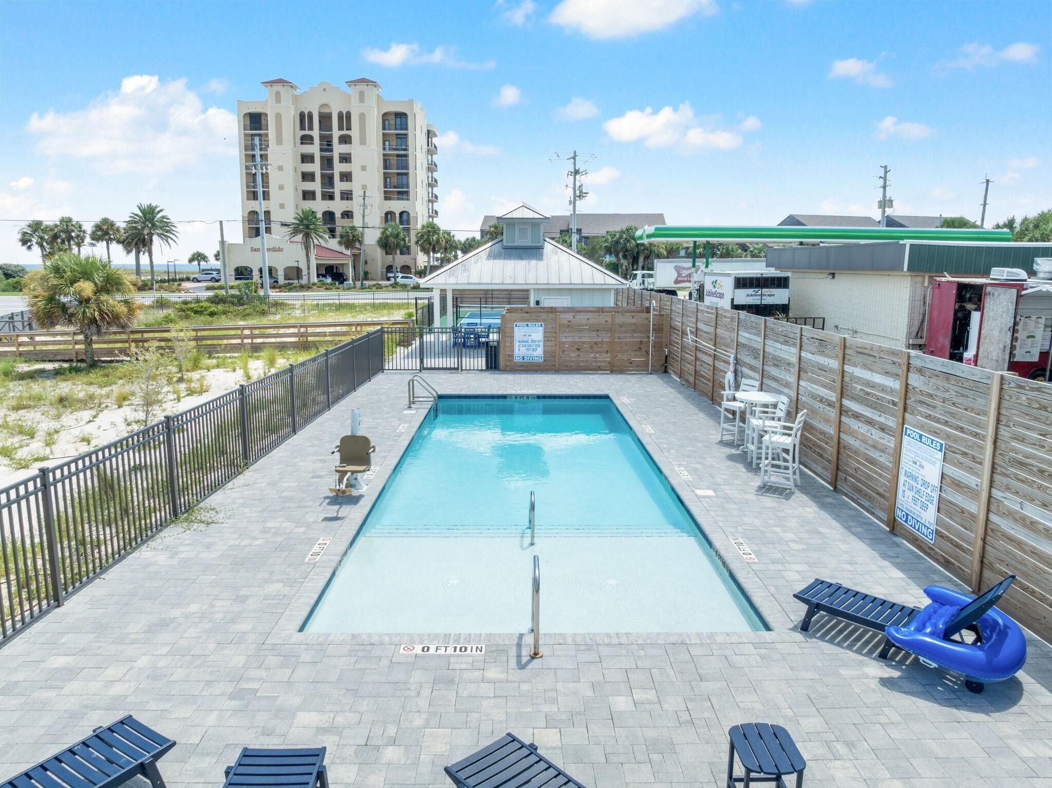 A rectangular pool with a diving board, surrounded by a wooden fence, and a beach in the background.
