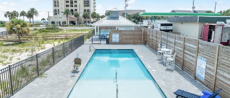 A rectangular pool with a diving board, surrounded by a wooden fence, and a beach in the background.