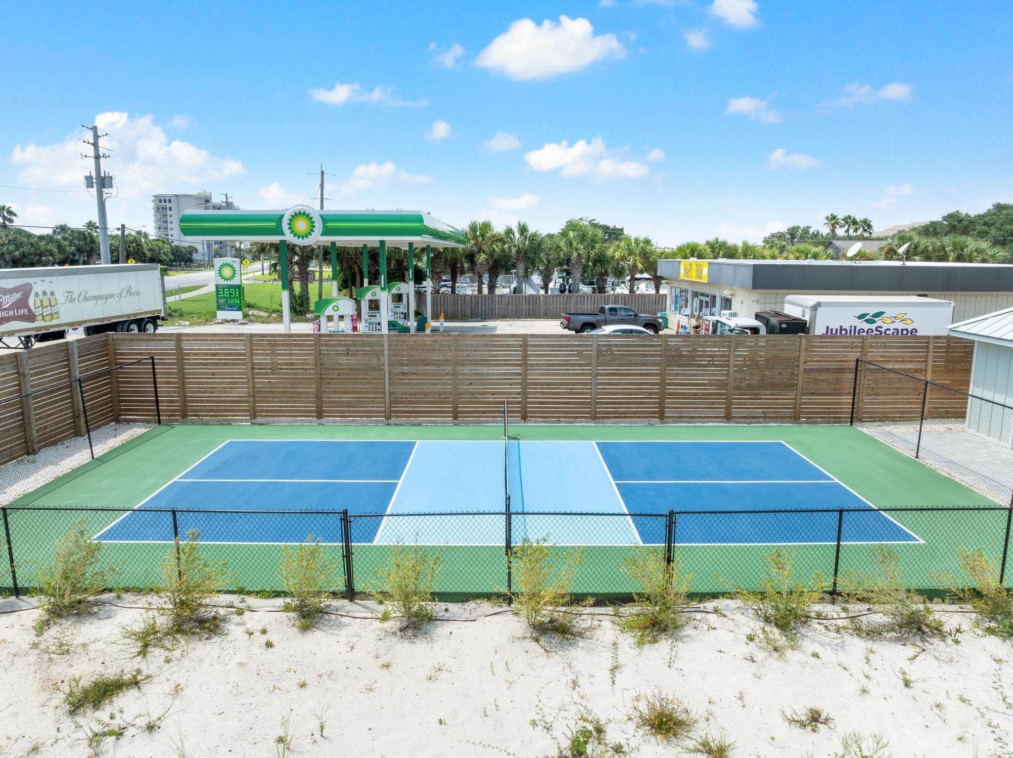 Ein Tennisplatz mit Zaun, Palmen und einer Tankstelle im Hintergrund.