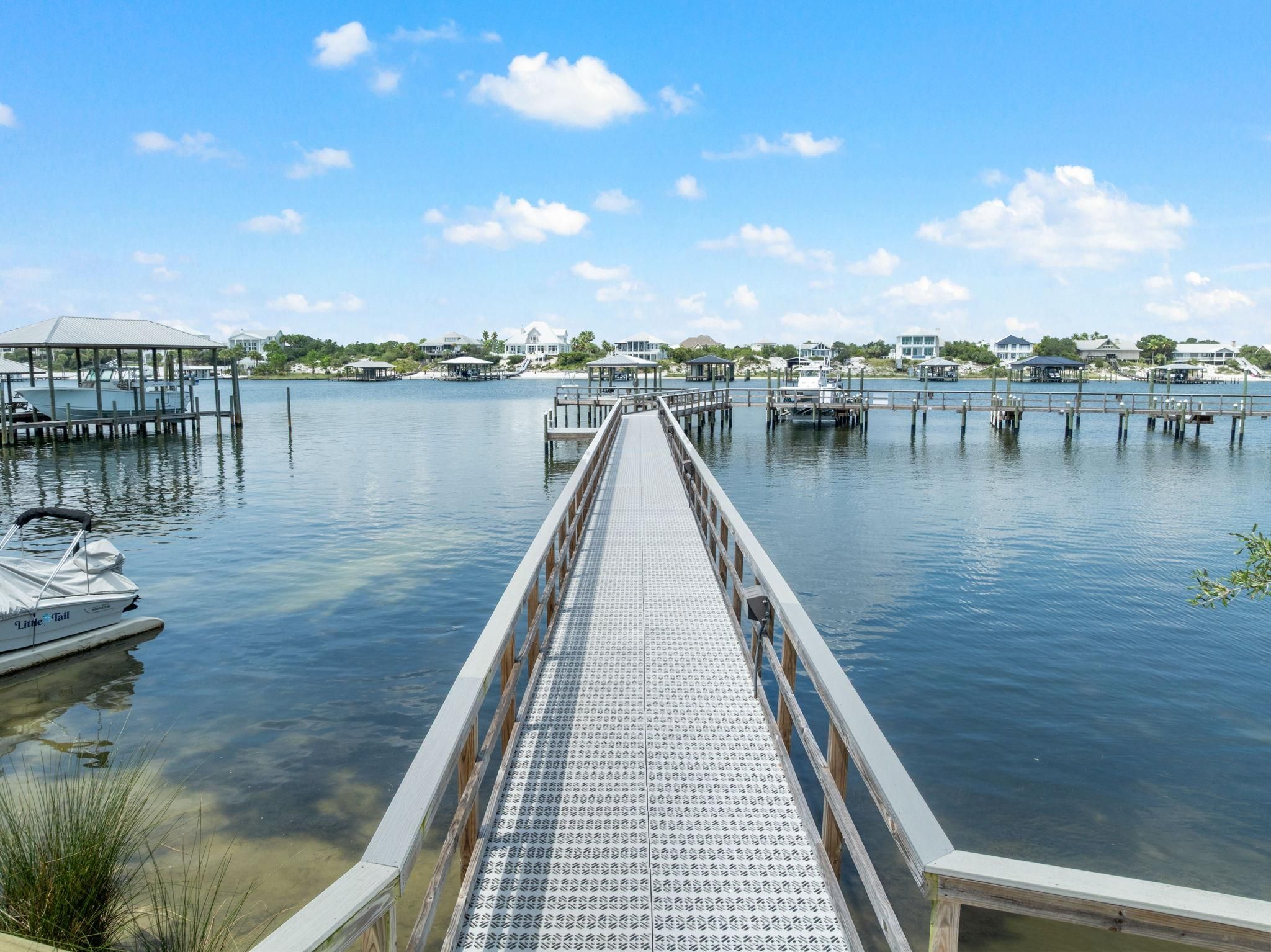 A pier leading to a marina with boats docked, surrounded by calm water and houses in the background.