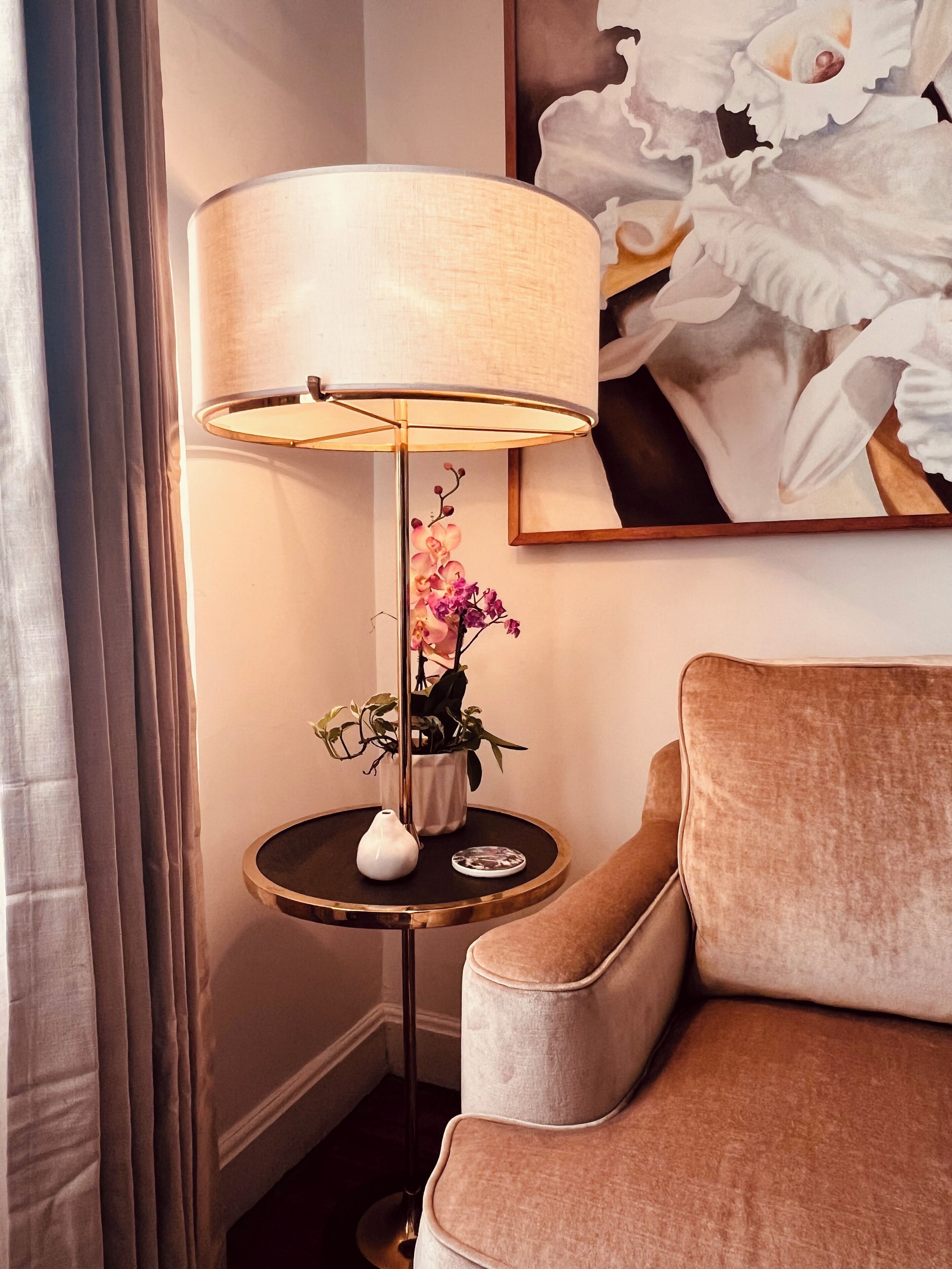 A room with a round table, a lamp, a vase with flowers, and a painting on the wall.