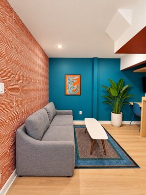 A modern living room with a grey sofa, a blue accent wall, a framed artwork, and a potted plant. - 1BR + Sofa Bed | Trendy Montrose (Houston)