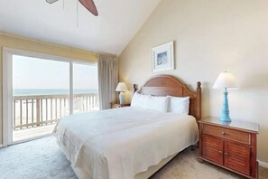 A hotel room with a large bed, a wooden headboard, two bedside lamps, a nightstand, a framed picture on the wall, and a view of the ocean through a sliding door. - Sweet Alabama (Gulf Shores)