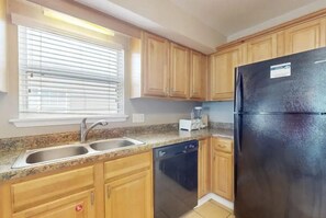 A kitchen with wooden cabinets, a black refrigerator, a dishwasher, and a window with blinds. - Sweet Alabama (Gulf Shores)