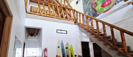 A multi-level interior with a checkered floor, a staircase with wooden railings, and surfboards leaning against the wall.