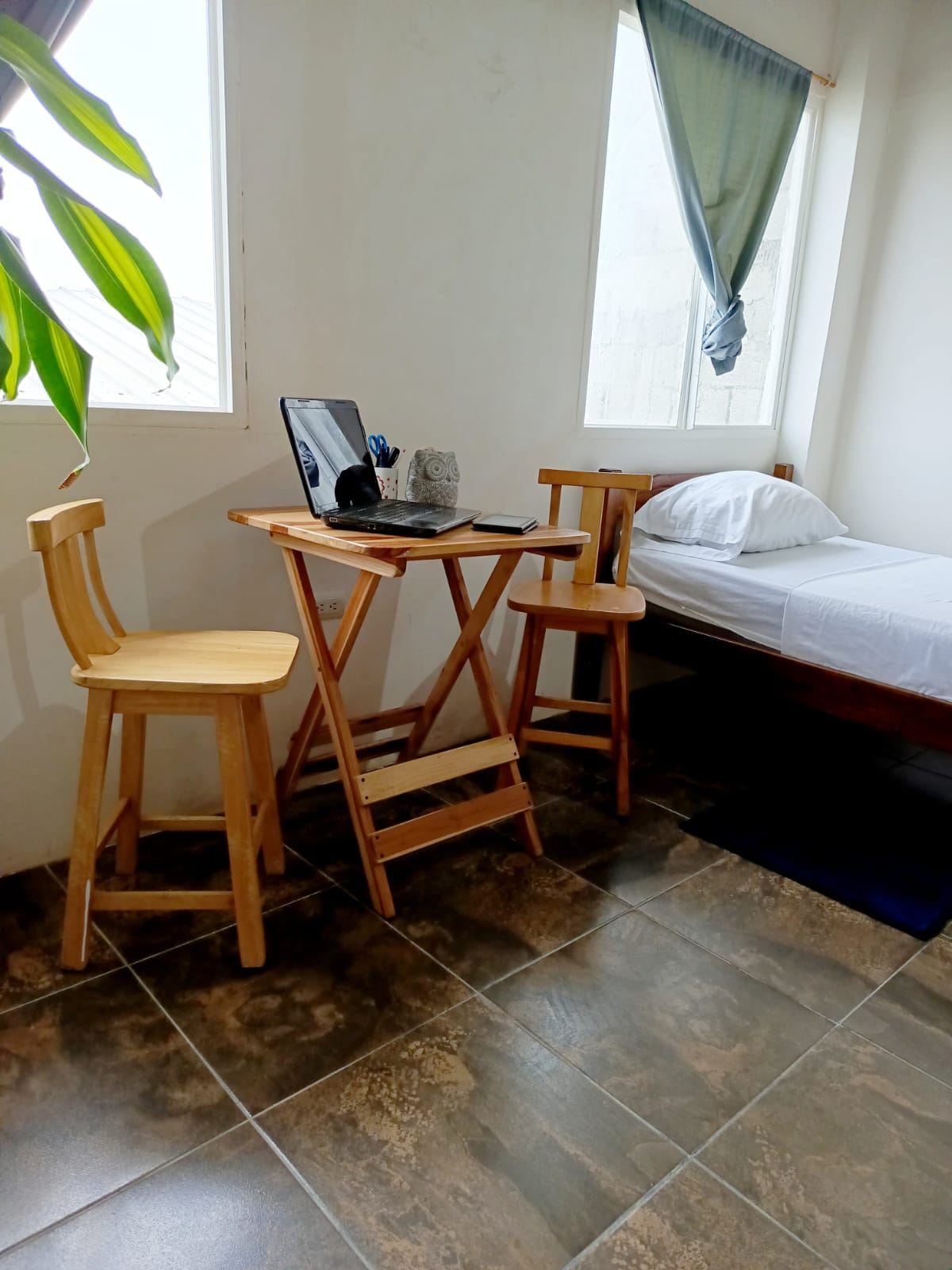 Ein kleines Zimmer mit einem Bett, einem Schreibtisch mit einem Laptop, zwei Stühlen und einem Fenster mit Vorhängen.