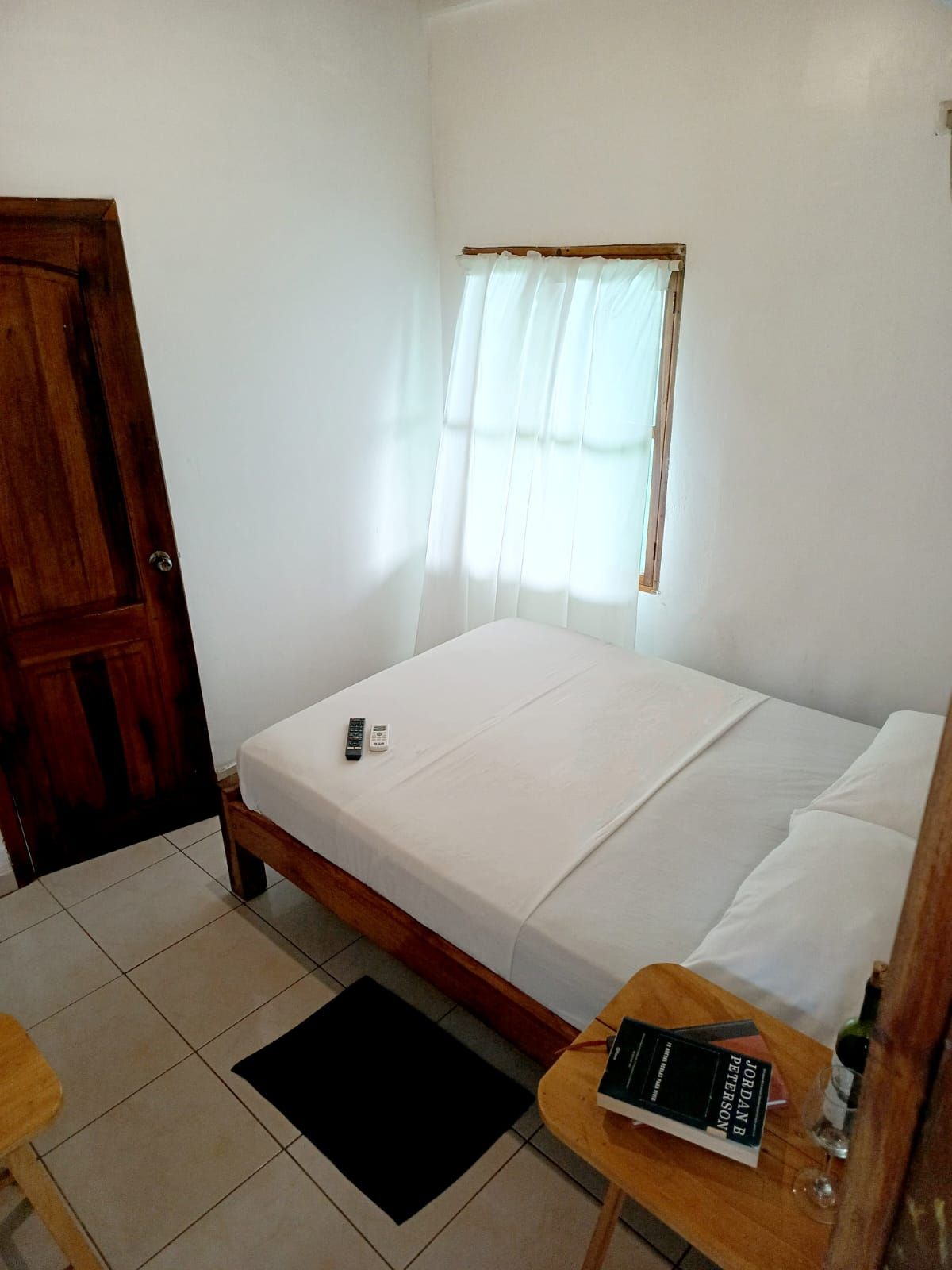 A single bed with a white mattress and pillows, a wooden door, a window with sheer curtains, a small table with a book and a bottle, and a black rug on the floor.