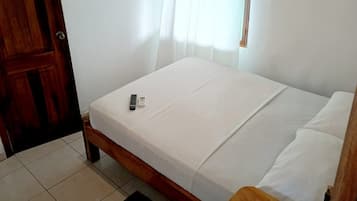 A single bed with a white mattress and pillows, a wooden door, a window with sheer curtains, a small table with a book and a bottle, and a black rug on the floor.