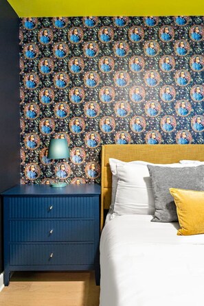 A bedroom with a patterned wallpaper, a wooden headboard, a bed with white and grey bedding, and a blue nightstand with a lamp. - Designer 1BR Suite | Trendy Montrose (Houston)