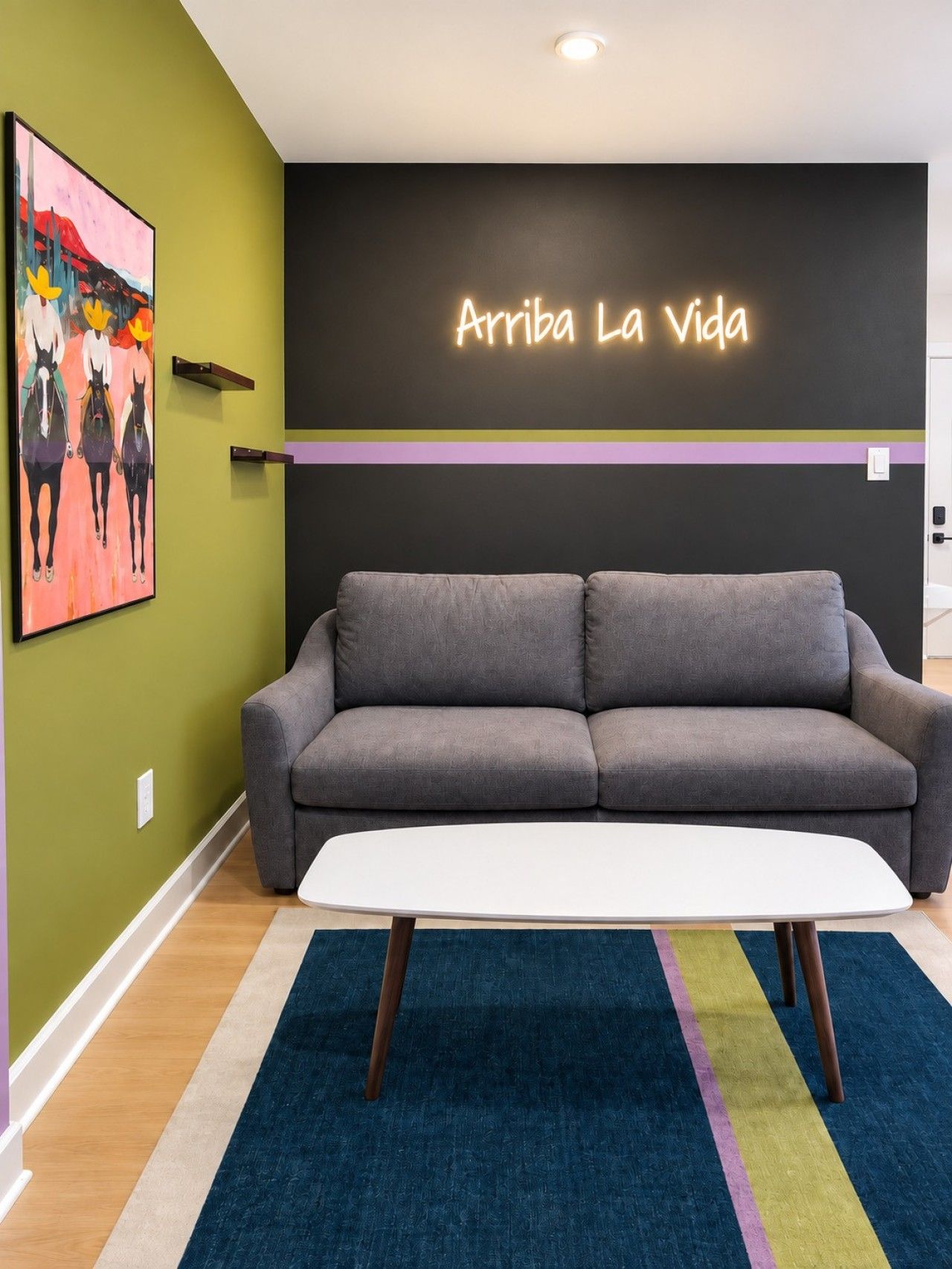 A room with a grey sofa, a white coffee table, a colorful rug, a wall with a neon sign, and a painting of horses.