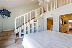 A bedroom with a bed, a staircase with white railings, a TV mounted on the wall, and a bathroom visible through an open door. - The Breeze (Gulf Shores)