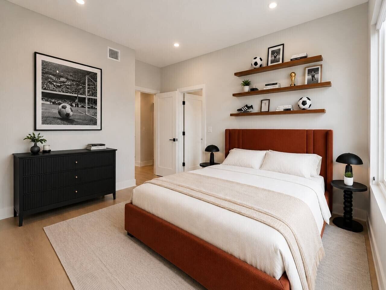 Un dormitorio con cama, cómoda, mesita de noche, lámpara, estante con libros y una pelota de fútbol, y una foto enmarcada en la pared.