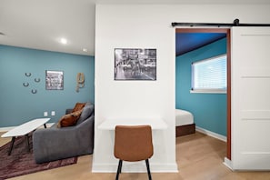 A modern room with a grey sofa, a white desk, and a brown chair. There is a black and white photo on the wall, a rope on the wall, and a window with blinds. - Trendy 1BR | Sleeps 4 | Free Parking (Houston)