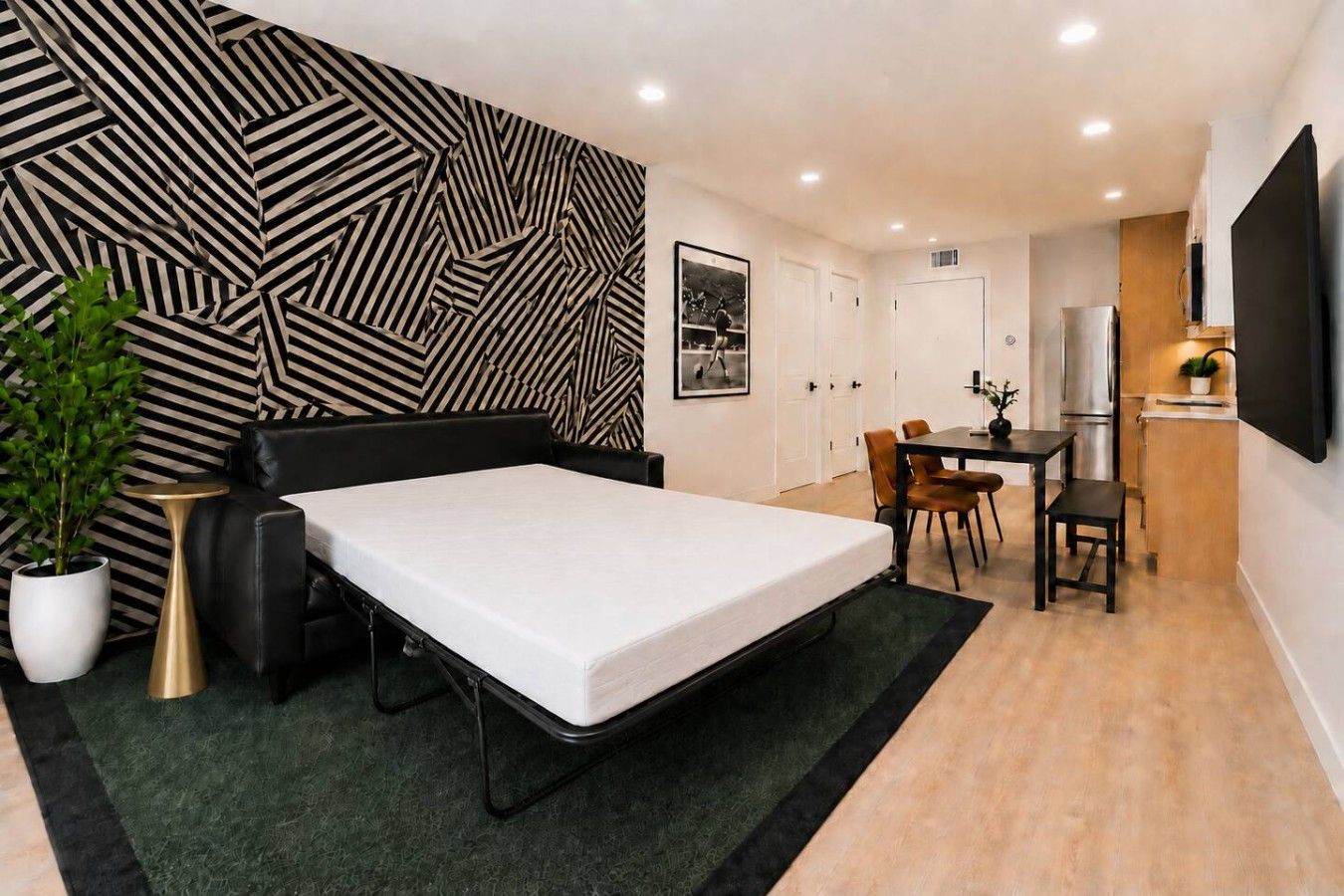 A compact room with a bed, a dining table, chairs, a small kitchen area, and a large TV.