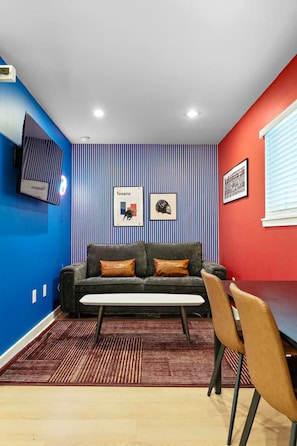 A room with a blue and red wall, a gray sofa with orange cushions, a wooden floor, and a TV mounted on the wall. - Trendy 1BR | DT Houston | Sleeps 4 (Houston)