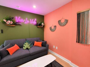 A living room with a sofa, pillows, a neon sign, wall decorations, and a curtain. - Modern 1BR | Free Parking | Sleeps 4 (Houston)