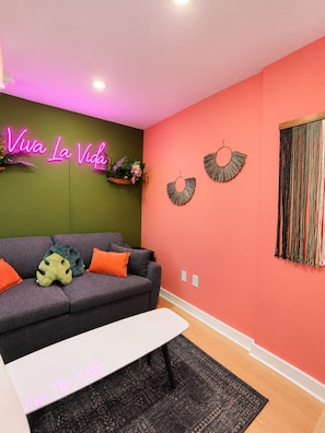 A modern living room with a sofa, cushions, a neon sign, wall art, and a patterned rug. - Modern 1BR | Free Parking | Sleeps 4 (Houston)