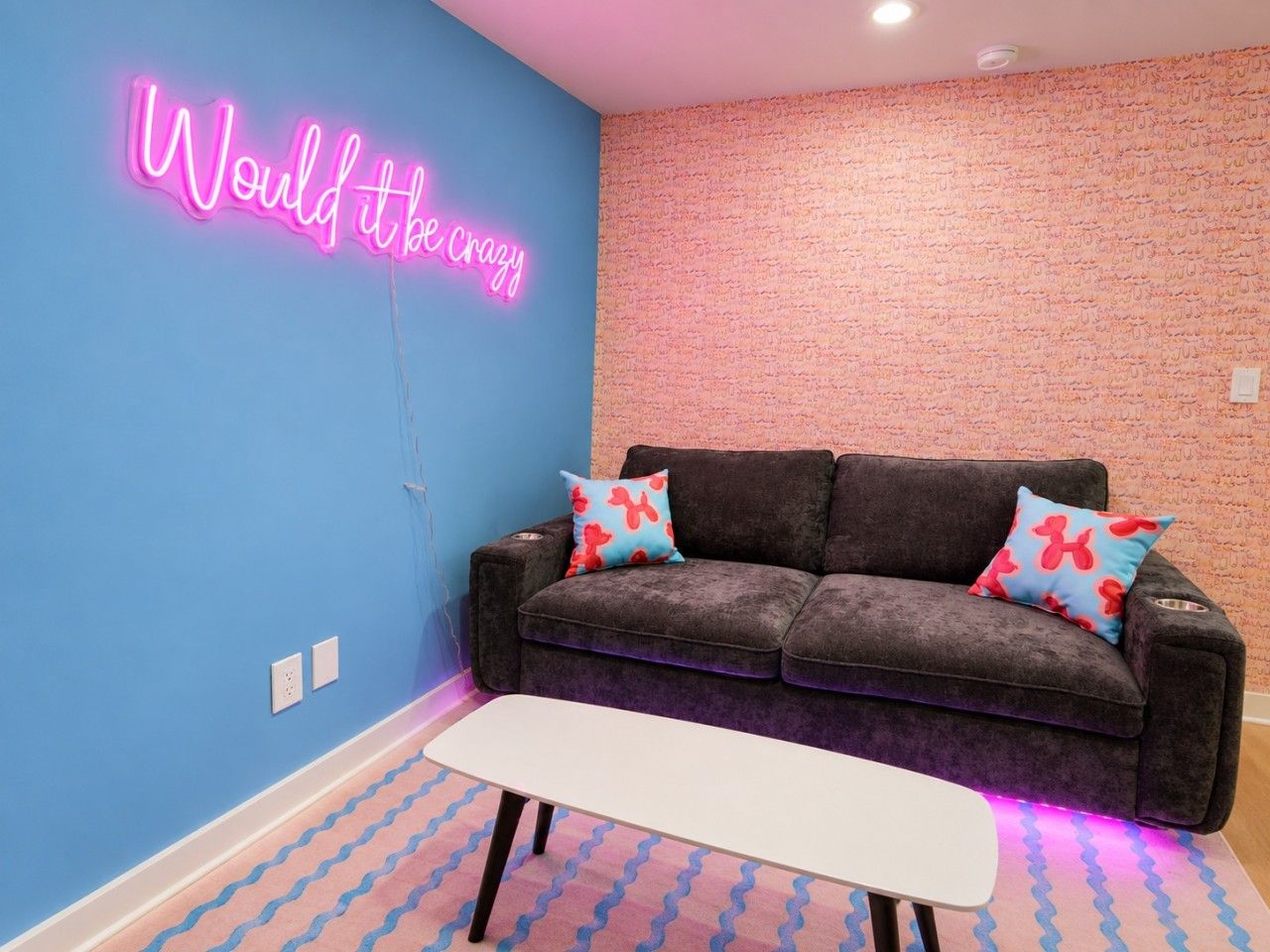 A room with a blue wall, a neon sign, a grey sofa with patterned cushions, and a white table.