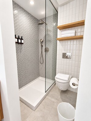 Ein modernes Badezimmer mit einer gläsernen Duschkabine, weiß gefliesten Wänden, einer Toilette und einem hölzernen Ablagebrett mit Handtüchern.