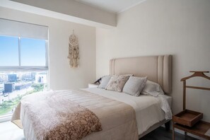 A bedroom with a large bed, a bedside table, a hanging dreamcatcher, and a view of the city through a window. - La Buenavista Prado Residences (La Victoria)