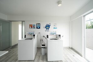Une buanderie équipée de deux machines à laver, d’une poubelle et de panneaux d’information accrochés au mur.