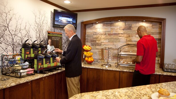 A breakfast bar with coffee machines, a fruit display, and a stone wall backdrop. - Accessible 1BR Suite w/ Kitchen & Hot Breakfast (Johnson City)