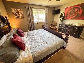 Ein Schlafzimmer mit einem großen Bett, einem Schminktisch, einem Sessel, einem Fernseher und einem Fenster mit Vorhängen.