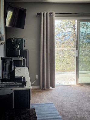 Ein Zimmer mit Fernseher, einer Kaffeemaschine und einer Pflanze auf einer Ablagefläche. Es gibt eine Glastür mit Vorhängen.